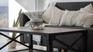 A modern living room with a dark grey sofa, white cushions, and a white bowl on a metal stand on a wooden table.