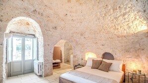 Une chambre avec un mur en pierre, un lit avec une literie blanche, une table de chevet avec une lampe et une porte donnant sur une autre pièce.