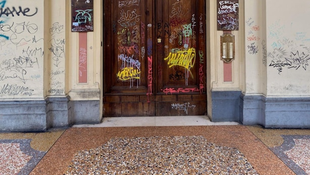 Entrada d'un edifici amb una porta doble de fusta, un sòl de rajola i un senyal al mur amb el número 17.