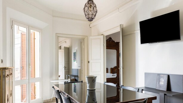 A dining area with a glass-top table, chairs, a chandelier, a flat-screen TV, and a view of the outside. - Irnerio Apts - Black Widow by Wonderful Italy (Bologna)