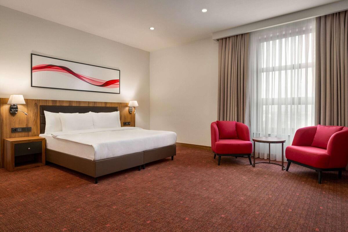 Une chambre d’hôtel avec un grand lit, deux tables de chevet avec des lampes, un fauteuil rouge, un canapé rouge, une petite table et une grande fenêtre avec des rideaux.