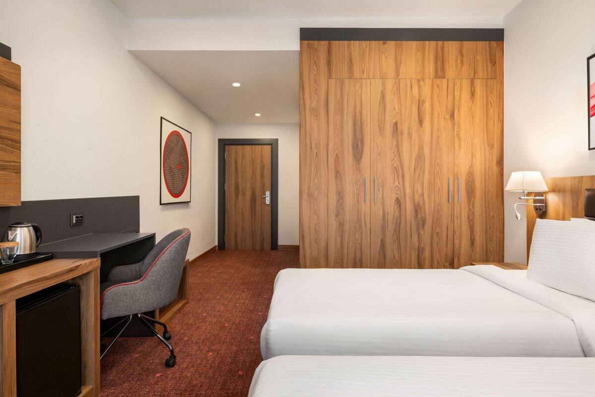 Une chambre d’hôtel équipée d’un bureau, de deux lits, d’une armoire en bois, d’un tableau encadré et d’une lampe.