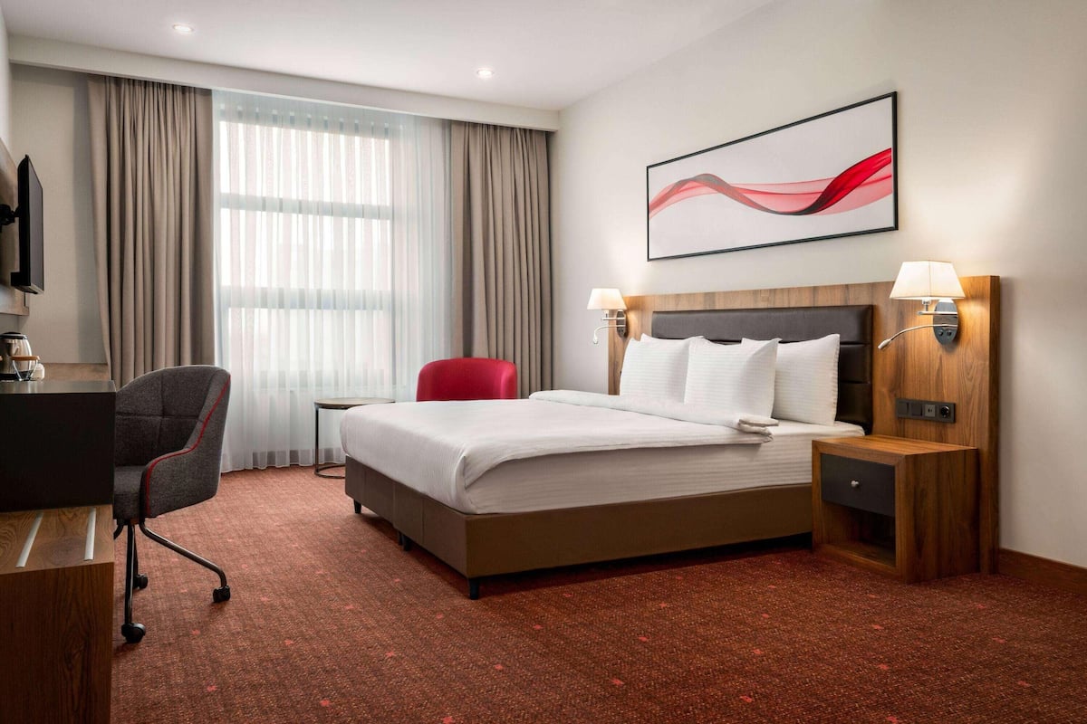 Une chambre d’hôtel avec un grand lit, deux tables de chevet avec des lampes, un bureau avec une chaise et une grande fenêtre avec des rideaux.
