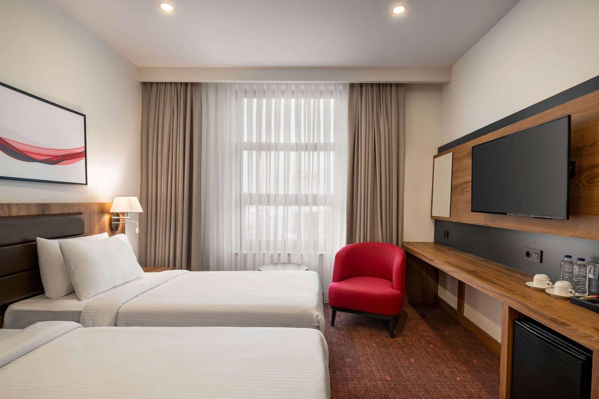 Une chambre d’hôtel avec deux lits, un fauteuil rouge, une télévision et une fenêtre avec des rideaux.