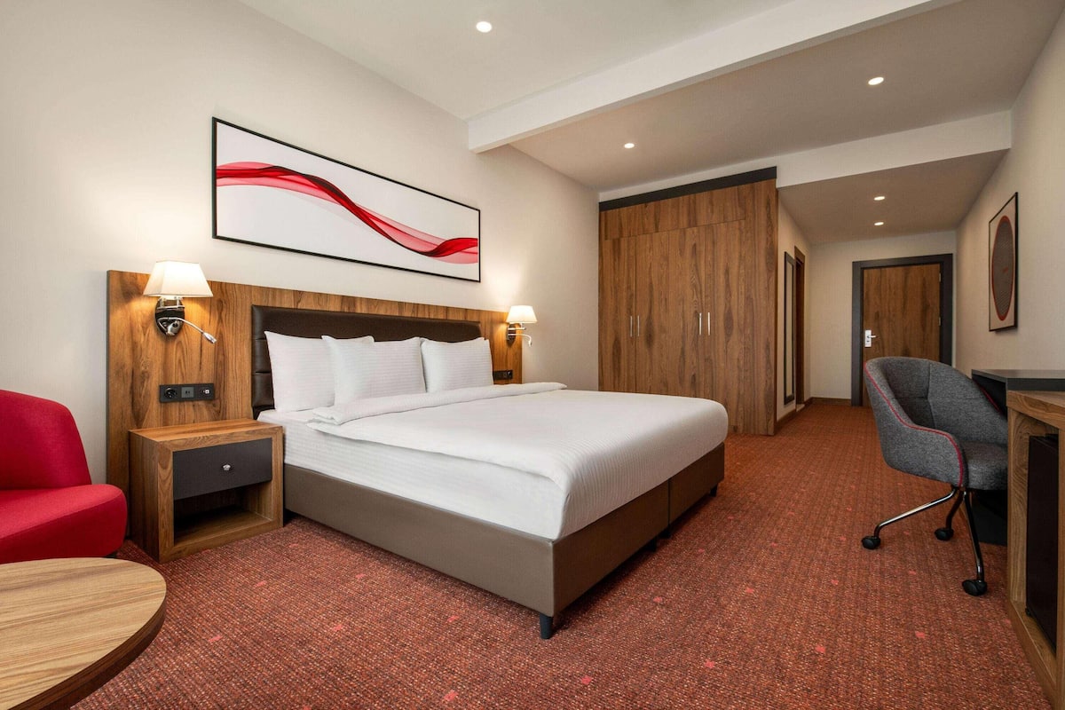 Une chambre d’hôtel avec un lit, deux tables de chevet avec des lampes, un fauteuil rouge, un bureau et un grand tableau abstrait accroché au mur.