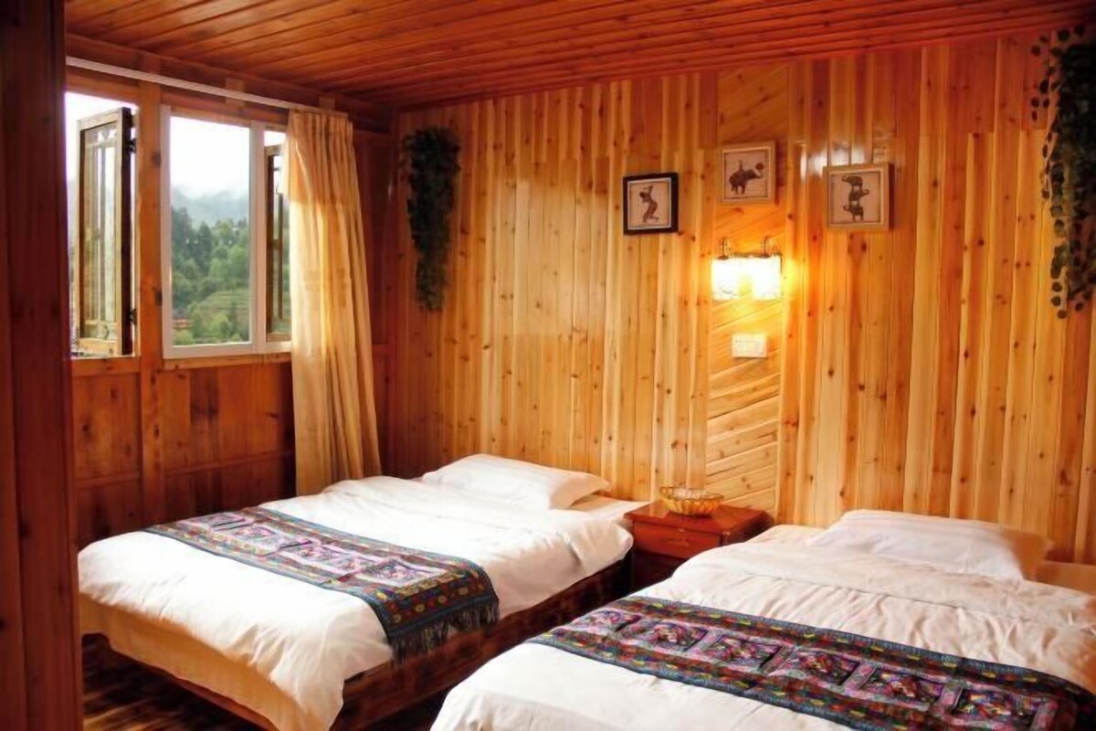 A wooden cabin room with two beds, a window with curtains, and wall-mounted lights.