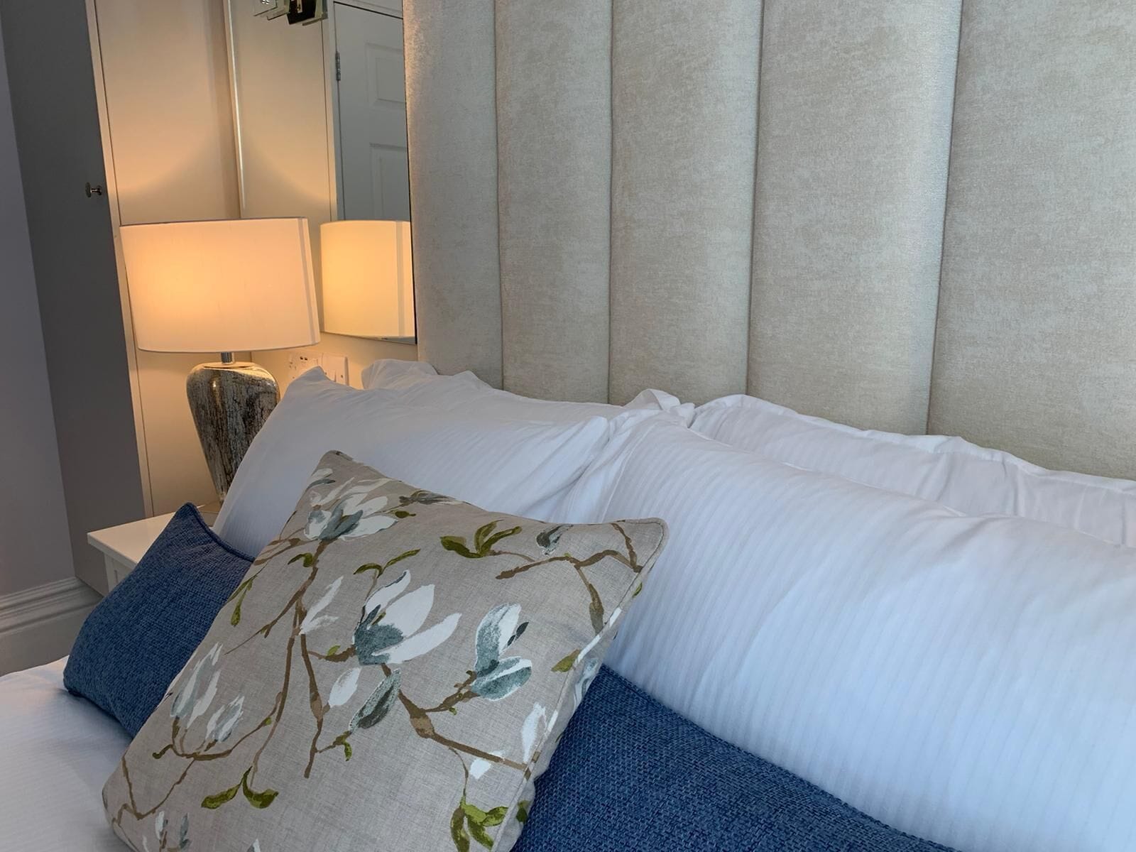 Una cama bien hecha con ropa de cama blanca y azul, un cojín con motivos florales y un cabecero con paneles verticales.