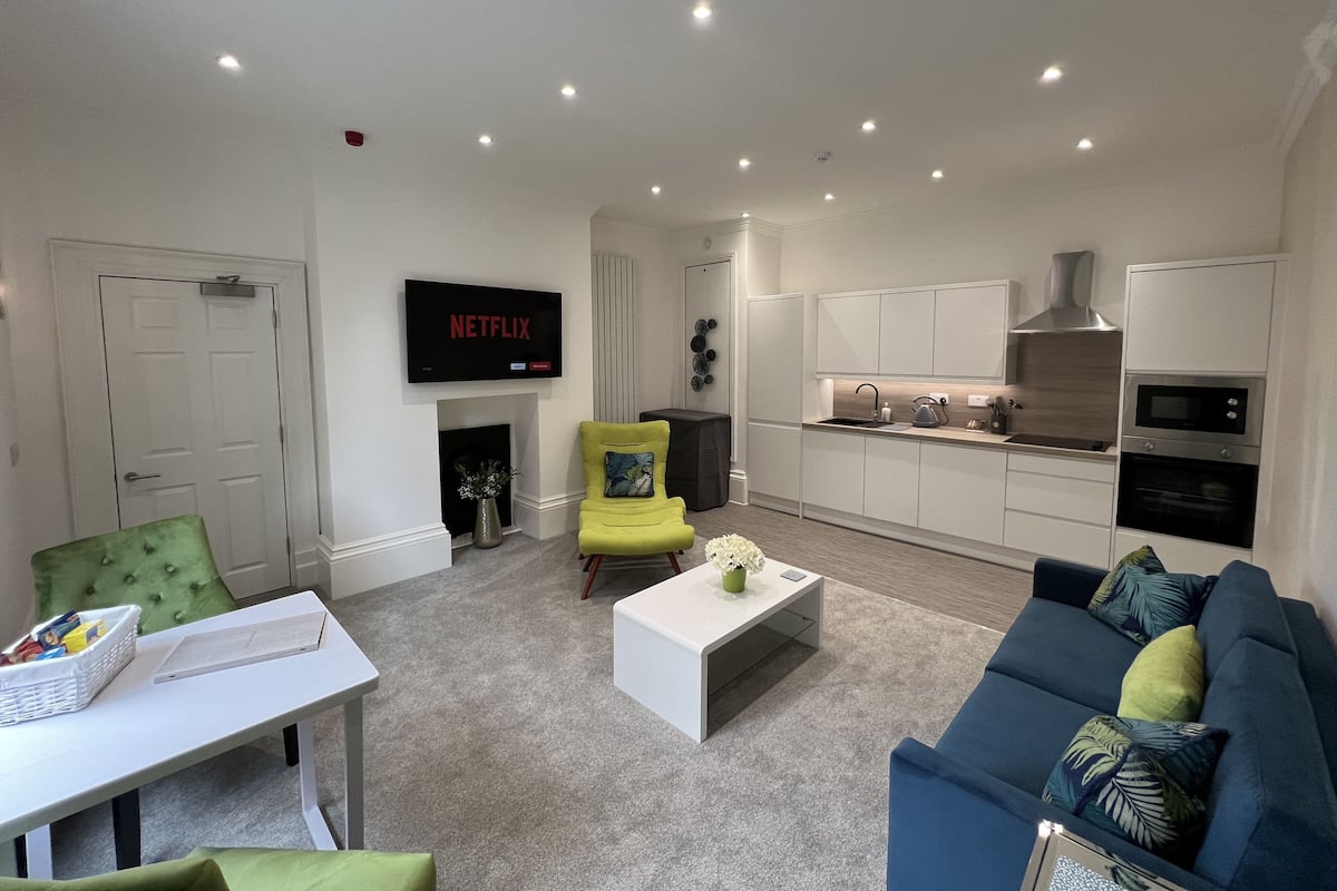 A modern living room with a sofa, a coffee table, a TV mounted above a fireplace, and a kitchen area with cabinets and appliances.