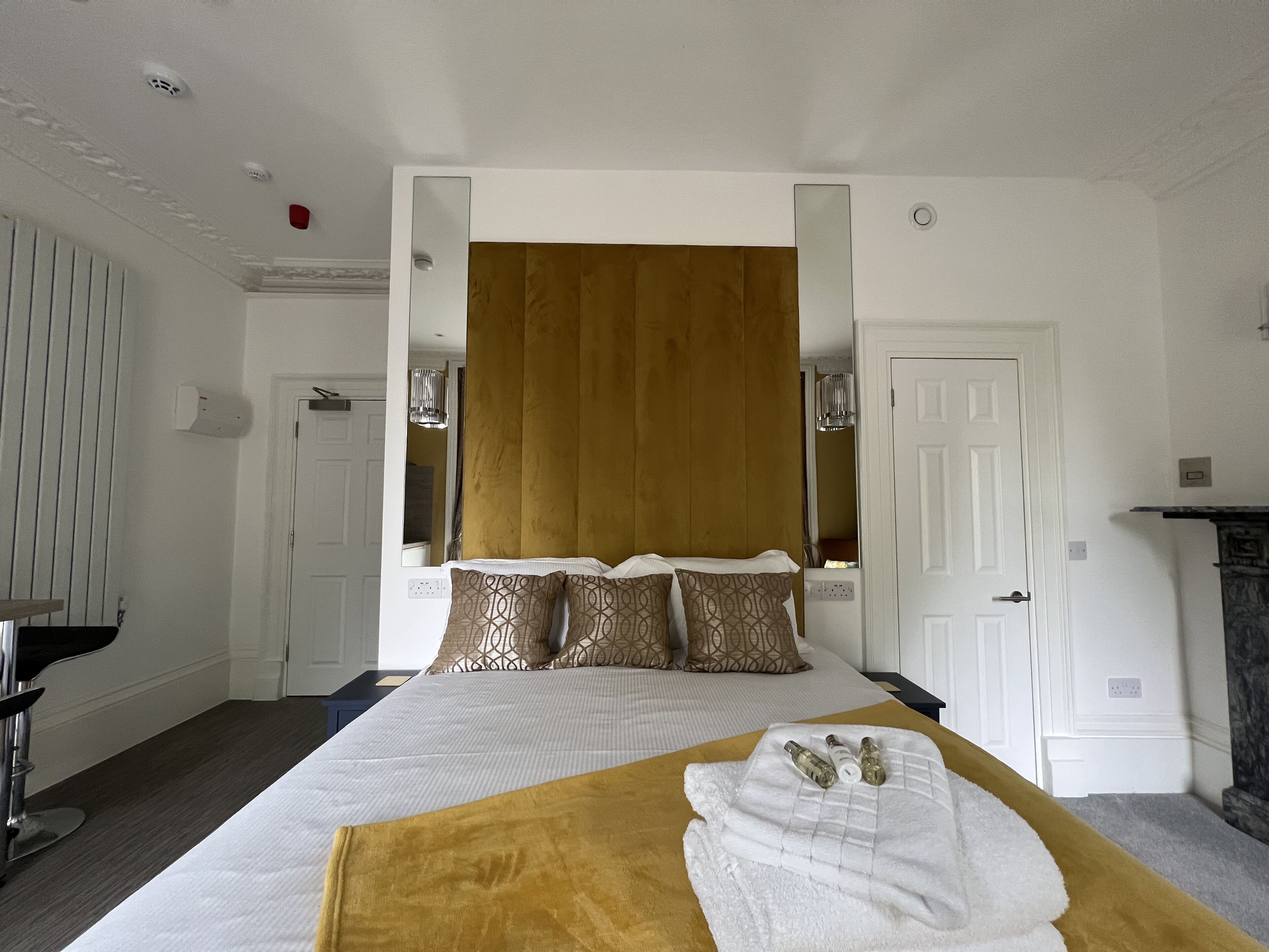 A hotel room with a large bed, a wooden headboard, a mirror, a white door, and a small table with a lamp.