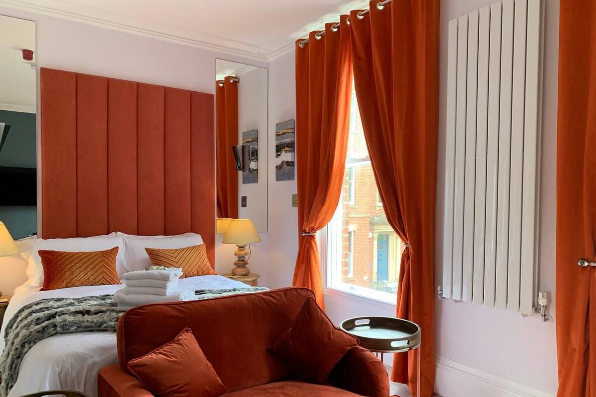 A hotel room with a bed, a red armchair, a side table, and a window with orange curtains.