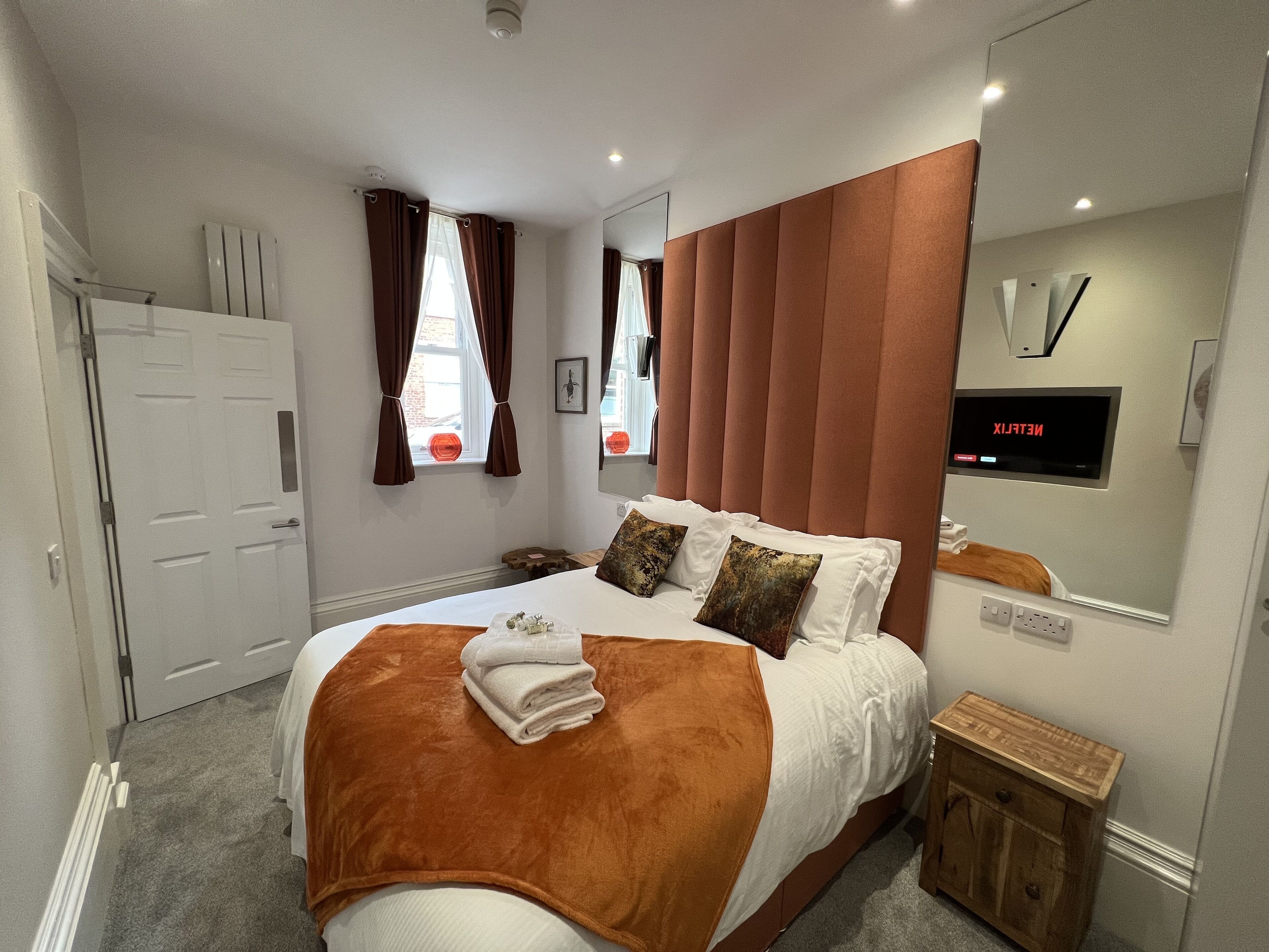 A modern bedroom with a large bed, a wooden nightstand, a mirror, and a wall-mounted TV displaying Netflix.