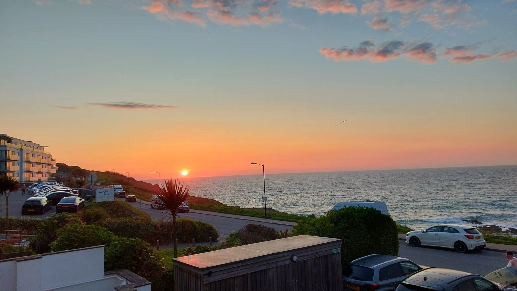 A coastal sunset view with parked cars and a building in the foreground.