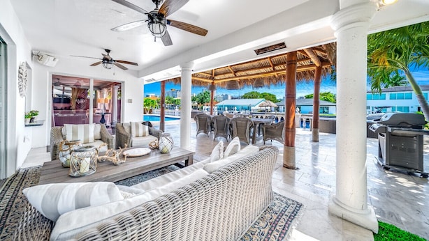 Un'ampia area lounge all'aperto con mobili in vimini, ventilatori a soffitto e vista su una piscina e edifici sullo sfondo.