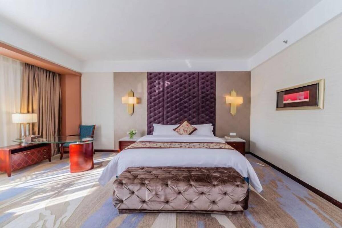 A hotel room with a large bed, a tufted headboard, a glass coffee table, a red ottoman, a desk with a lamp, and a framed picture on the wall.