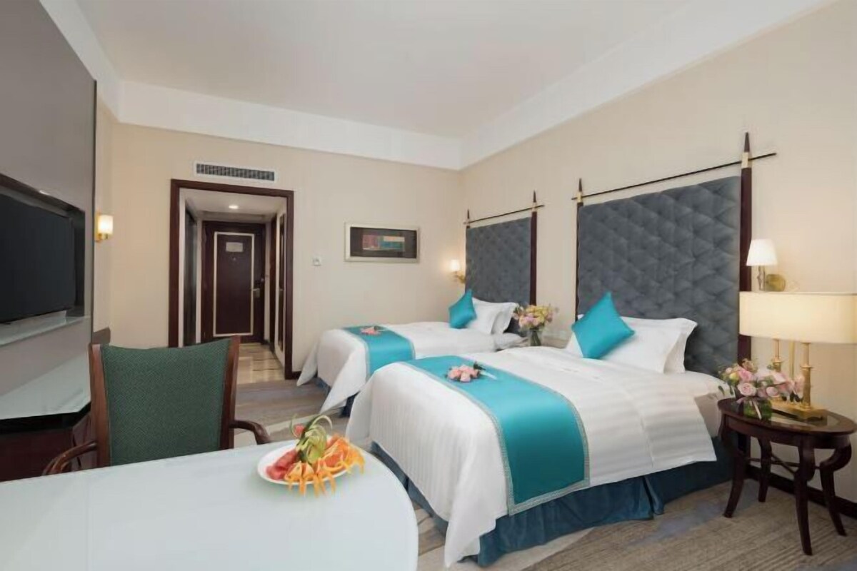 A hotel room with two beds, a TV, a dining table with fruit, and a small table with a lamp.