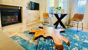 Een moderne woonkamer met een open haard, een flatscreen televisie aan de muur, een eettafel met twee stoelen en een grote houten salontafel.