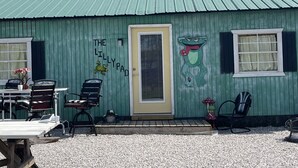 Un bâtiment à l’extérieur vert, avec une porte jaune et une fenêtre dotée de volets blancs. On y trouve deux chaises et une table à l’extérieur.