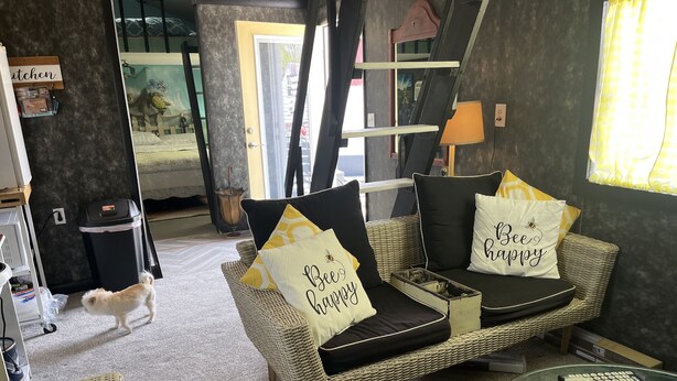 Un salon chaleureux avec un canapé en osier, deux coussins ornés de la mention « Soyez heureux », une table basse en verre et un petit chien.