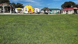 Un champ herbeux avec plusieurs petits bâtiments, dont une structure jaune et rouge, un bâtiment surmonté d’un toit vert et un bâtiment blanc avec un toit rouge.