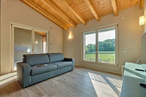 Une pièce avec un canapé gris, des poutres en bois au plafond, une grande fenêtre donnant sur un paysage verdoyant et un bureau blanc.