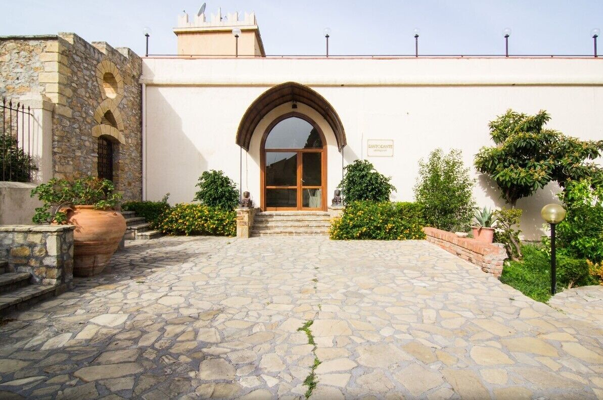 A stone-paved entrance with a large arched doorway, a white building with a sign, and a small garden area with potted plants.