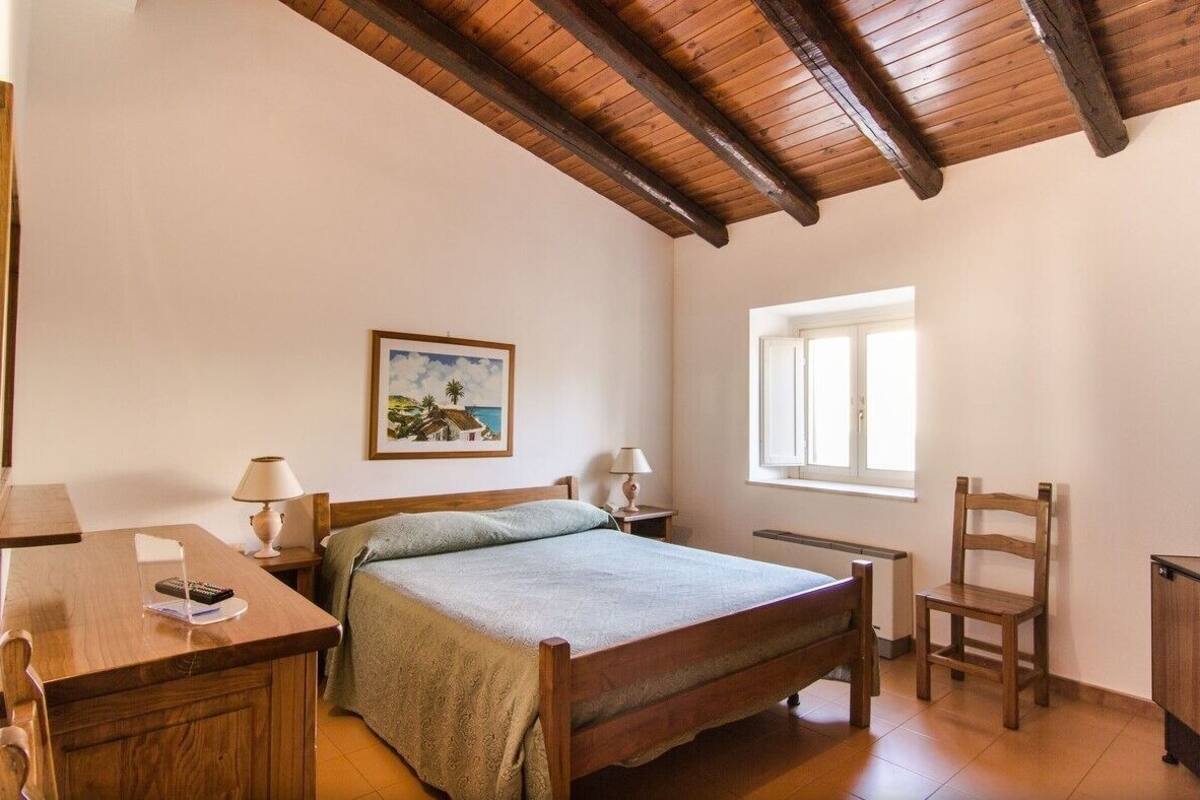 Un dormitorio con cama de madera, un escritorio con lámpara, una silla, un ventanal y un cuadro en la pared.