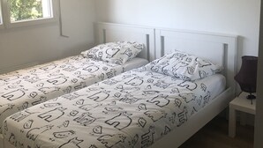 A bedroom with a white bed, a bedside table, a lamp, a hanging light fixture, and a window with a view of trees. - Villa 10 minutes from the quiet beach (Soulac-sur-Mer)