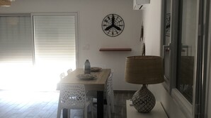 A modern kitchen with a wooden table, white chairs, a clock, and a window with blinds. - Villa 10 minutes from the quiet beach (Soulac-sur-Mer)