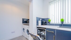 A compact kitchen with a washing machine, dishwasher, and a window with blinds. - The Annexe, 25 Gorsehill Road (Wirral)