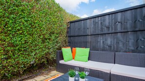 An outdoor seating area with a dark-colored sofa, green and orange cushions, and a small table with potted plants and wine glasses. - The Annexe, 25 Gorsehill Road (Wirral)