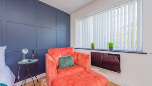 A modern bedroom with a large bed, a small side table, a bright orange sofa, a window with blinds, and a radiator. - The Annexe, 25 Gorsehill Road (Wirral)