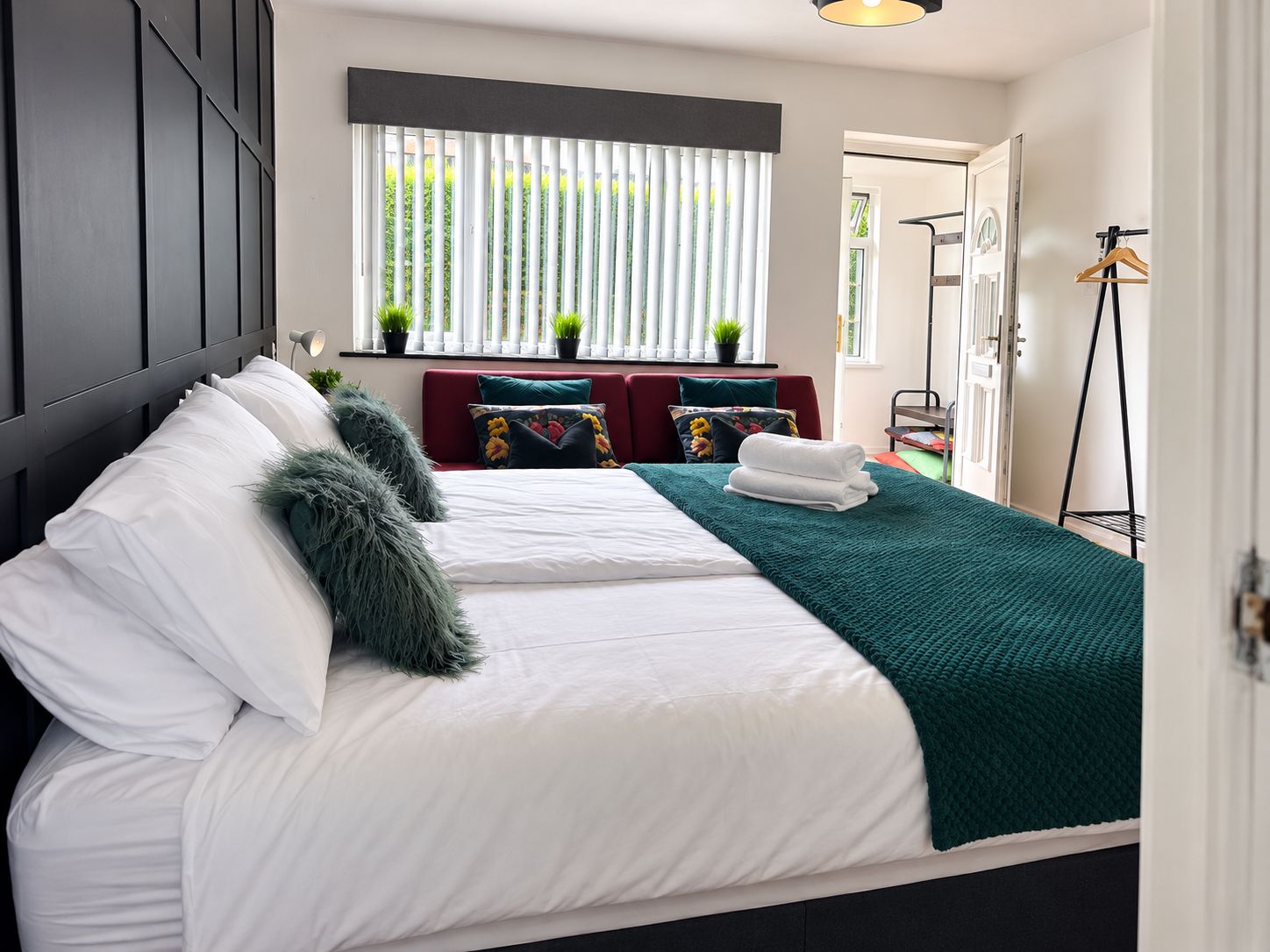 Un letto rifatto con lenzuola bianche e una coperta verde, un divano rosso con cuscini floreali e una finestra con persiane verticali.