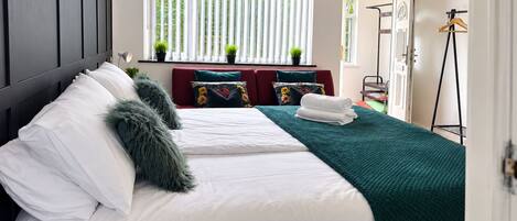 Una cama bien tendida con sábanas blancas y un plaid verde, un sofá rojo con cojines florales, y una ventana con persianas verticales.