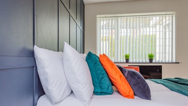 A neatly made bed with white and colorful pillows, a television on a stand, and a window with blinds. - The Annexe, 25 Gorsehill Road (Wirral)