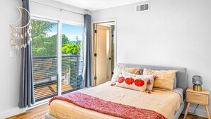 A bedroom with a bed, a nightstand, a lamp, a dreamcatcher, and a view of greenery through the window.
