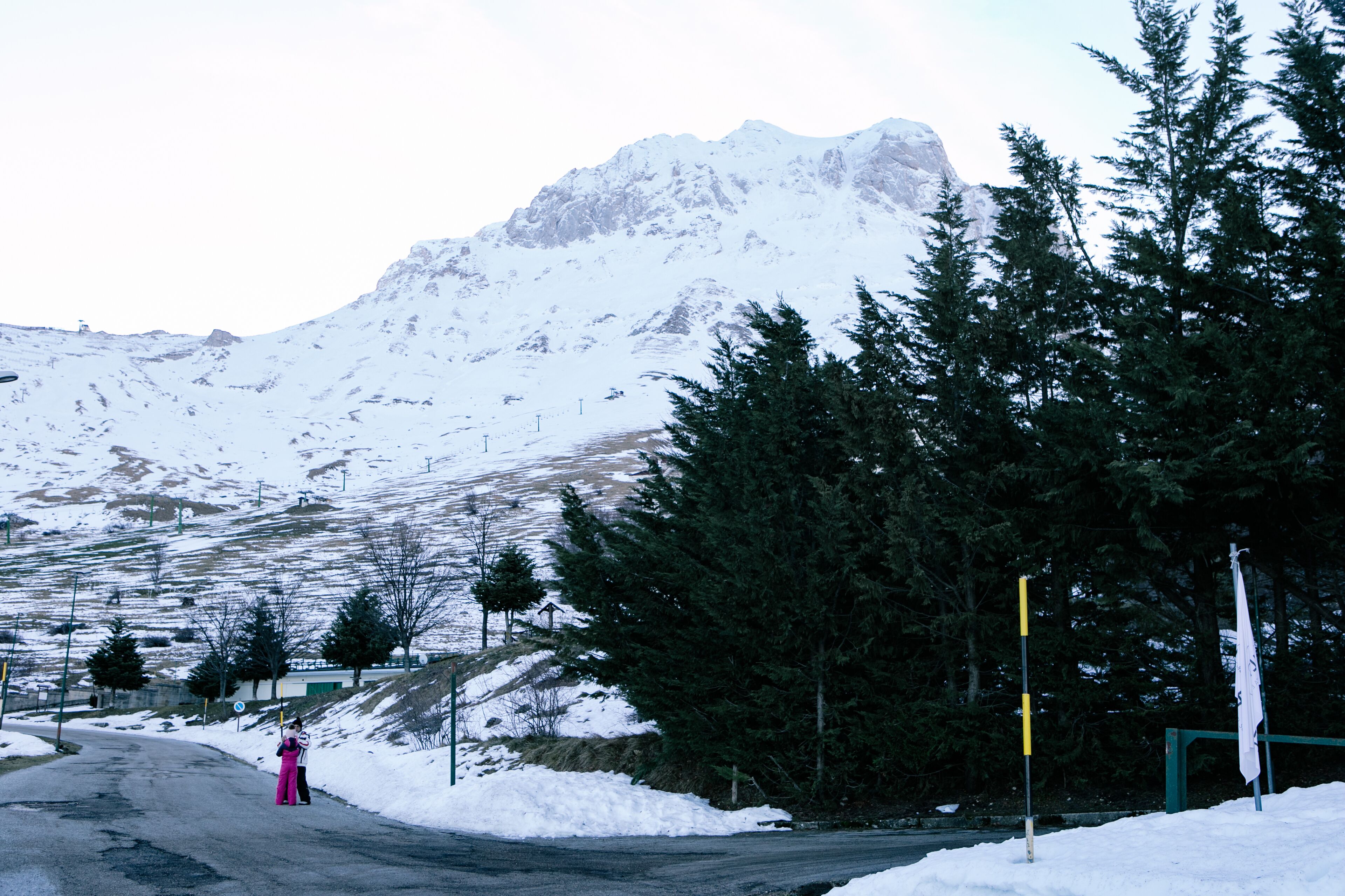 A snow-covered mountain road with a person in pink clothing standing by the roadside.