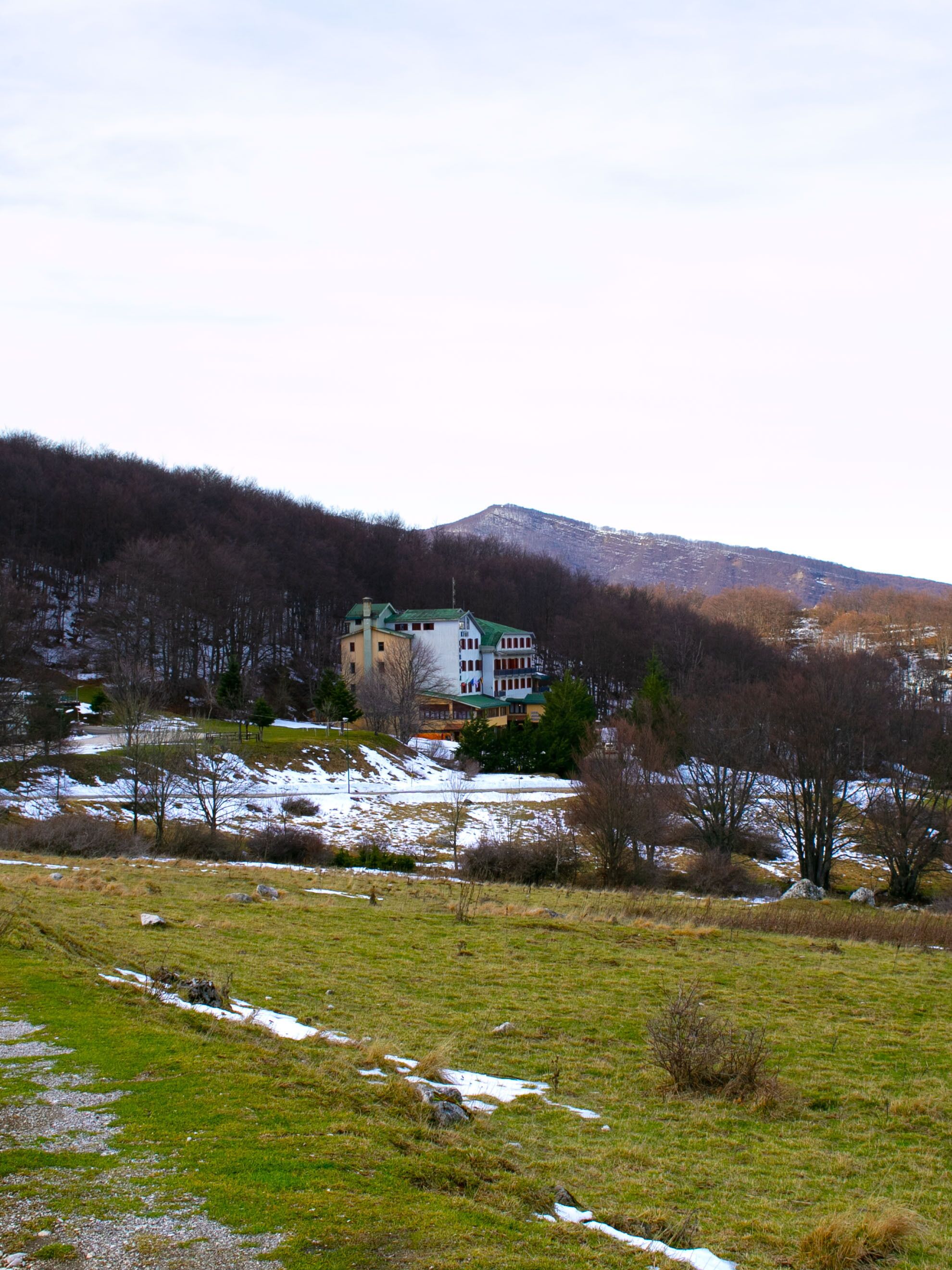 A building with a green roof and multiple stories, surrounded by a natural landscape with snow patches and trees.