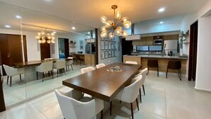 A modern dining area with a large wooden table and white chairs, a kitchen with a microwave and stove, and a hallway leading to a bedroom. - Moderno departamento con alberca, 200m al malecón (Mazatlán)