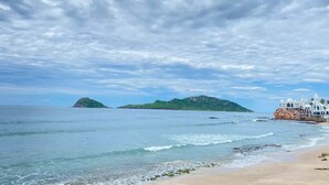 A beach with clear waters, a sandy shore, and a white building with a rocky base on the right. - Moderno departamento con alberca, 200m al malecón (Mazatlán)