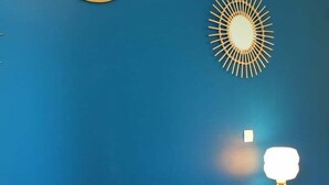A room with a blue wall, a round mirror, a sunburst wall decoration, a bedside table with a lamp, and patterned bedding.