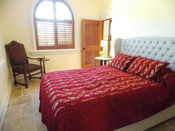 Un dormitorio con una cama grande, una silla de madera, una mesita y una ventana con persianas.