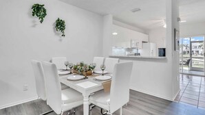 A modern dining room with a white table set for four, a kitchen in the background, and a view of the outdoors through a glass door. - Townhouse •Pool •Air Hockey• Gated •Near Beach &IMG • 3 beds (Bradenton)