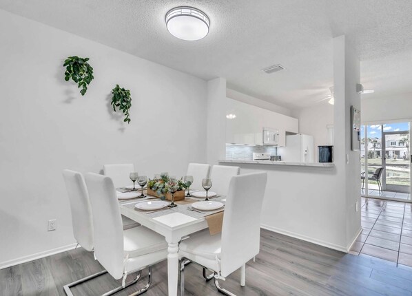 A modern dining room with a white table set for four, a kitchen in the background, and a view of the outdoors through a glass door. - Townhouse •Pool •Air Hockey• Gated •Near Beach &IMG • 3 beds (Bradenton)