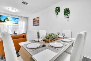 A dining area with a long table set for four, white chairs, and a view of the beach through a window. - Townhouse•Heated Pool•Lake view•Beach Stuff•3 beds• (Bradenton)