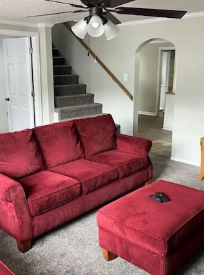 A red sofa and ottoman in a living room with a ceiling fan, carpeted floor, and a staircase in the background.