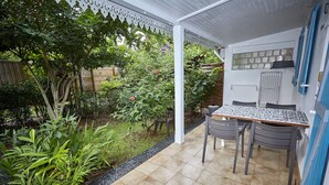 Un porche cubierto con piso de baldosas, una mesa con sillas y diversos plantas y árboles.