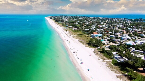 Eine Vogelperspektive einer Küstenregion mit einem langen Sandstrand, klarem türkisblauem Wasser und einer dicht bebauten Wohngegend mit Häusern und Gebäuden.