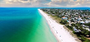 Vue aérienne d’une zone côtière avec une longue plage de sable, des eaux turquoise et limpides, et une zone résidentielle densément peuplée, avec des maisons et des immeubles.