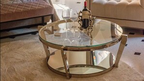 Quarto bem decorado com cama, mesa de centro redonda de vidro com copos de vinho, mesa lateral com garrafas decorativas, luminária e vista pela janela.
