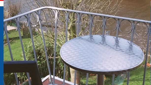En balkong med ett runt bord, med utsikt över en flod och en trädgård med träd och ett hus i bakgrunden.